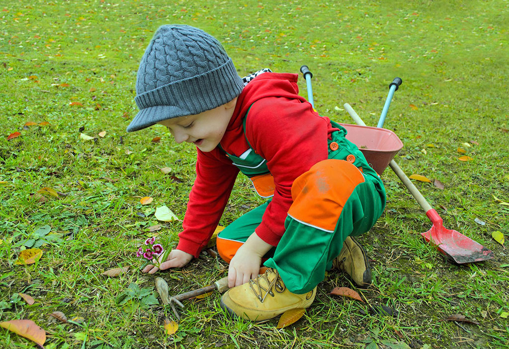Young boy in green overalls, a red hoodie, and a knit hat, gardening.