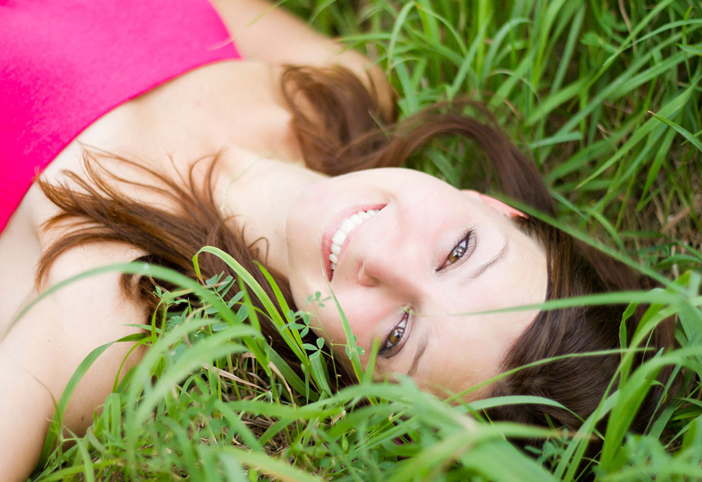 Brunette woman smiling, laying in tall green grass.